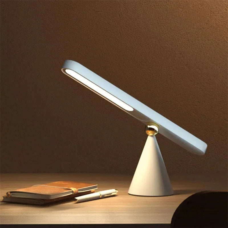 Geometric shaped white desk lamp with a magnetic base on a wooden table next to a book.