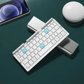 Silver white mini aluminium folding Bluetooth keyboard open next to a smartphone on its integrated stand.