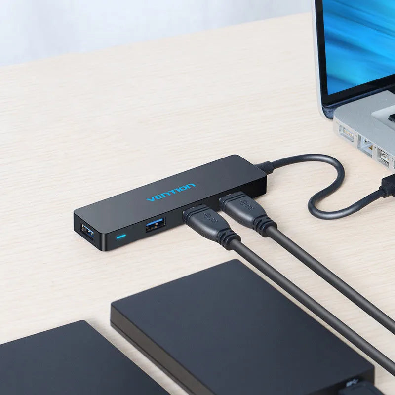 Black four-port USB 3.0 hub with a short cable on a desk next to a laptop.