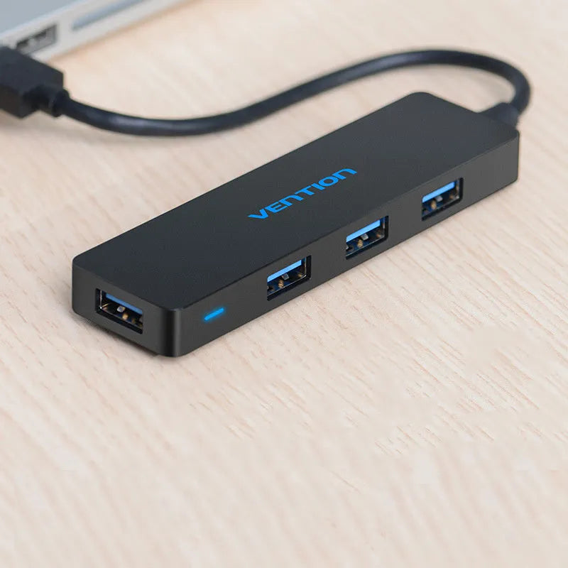 Black four-port USB 3.0 hub with a short cable on a desk next to a laptop.