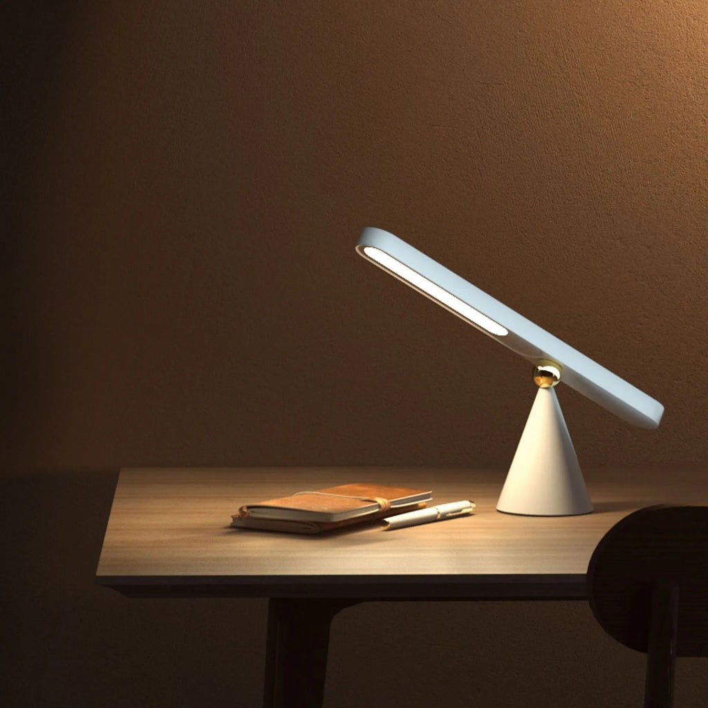 Geometric shaped white desk lamp with a magnetic base on a wooden table next to a book.