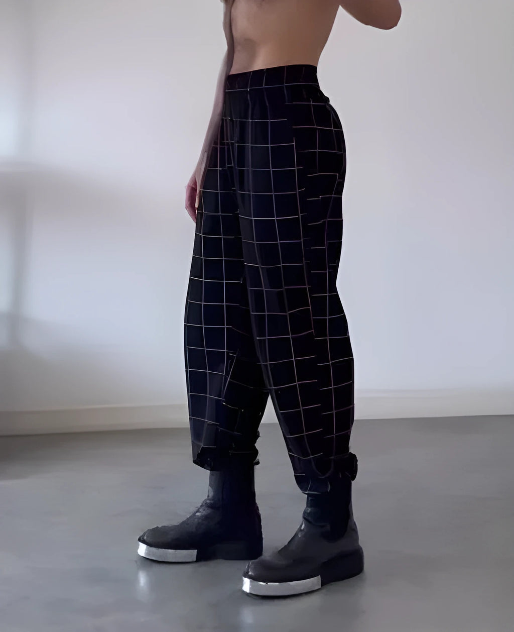 Man wearing grey plaid trousers with a distinctive apron-style waistband, showing the tartan pattern and zip fly.