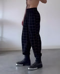 Man wearing grey plaid trousers with a distinctive apron-style waistband, showing the tartan pattern and zip fly.