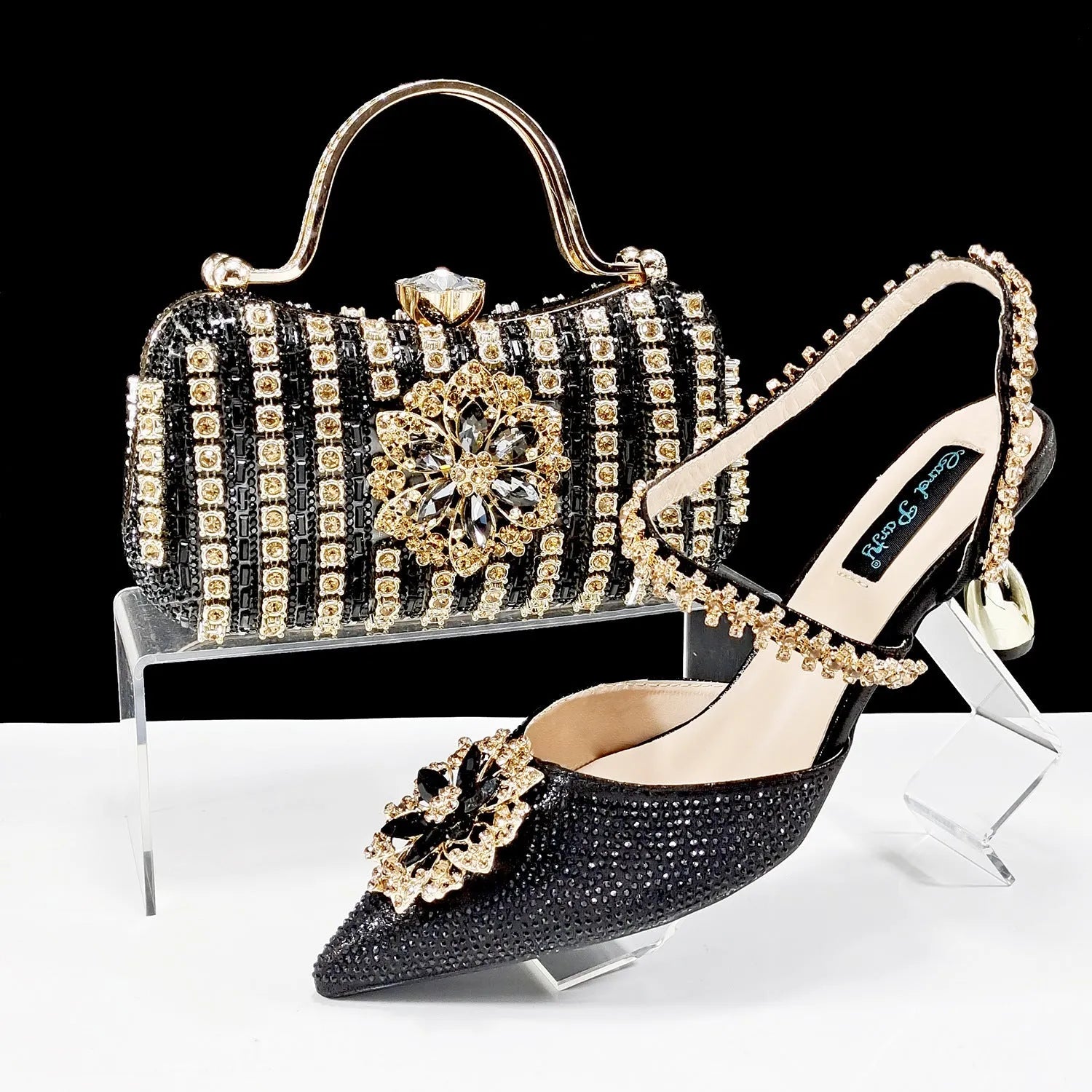 Women's black high heel ankle-strap sandals with floral buckle and chain detail next to matching clutch bag